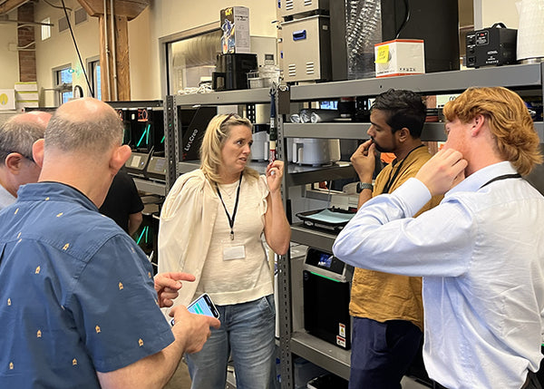 Group of people in a warehouse or storage area with shelves and equipment.